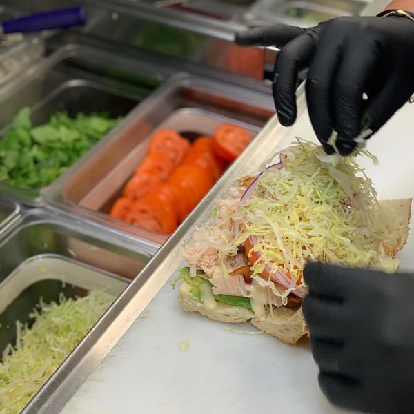 Gloved hands preparing a Taylor Gourmet sandwich, adding shredded lettuce over meats and vegetables.