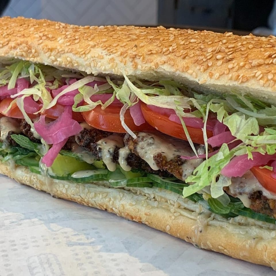 Taylor Gourmet sandwich with falafel, lettuce, tomatoes, pickled onions, and sauce on a sesame seed roll.